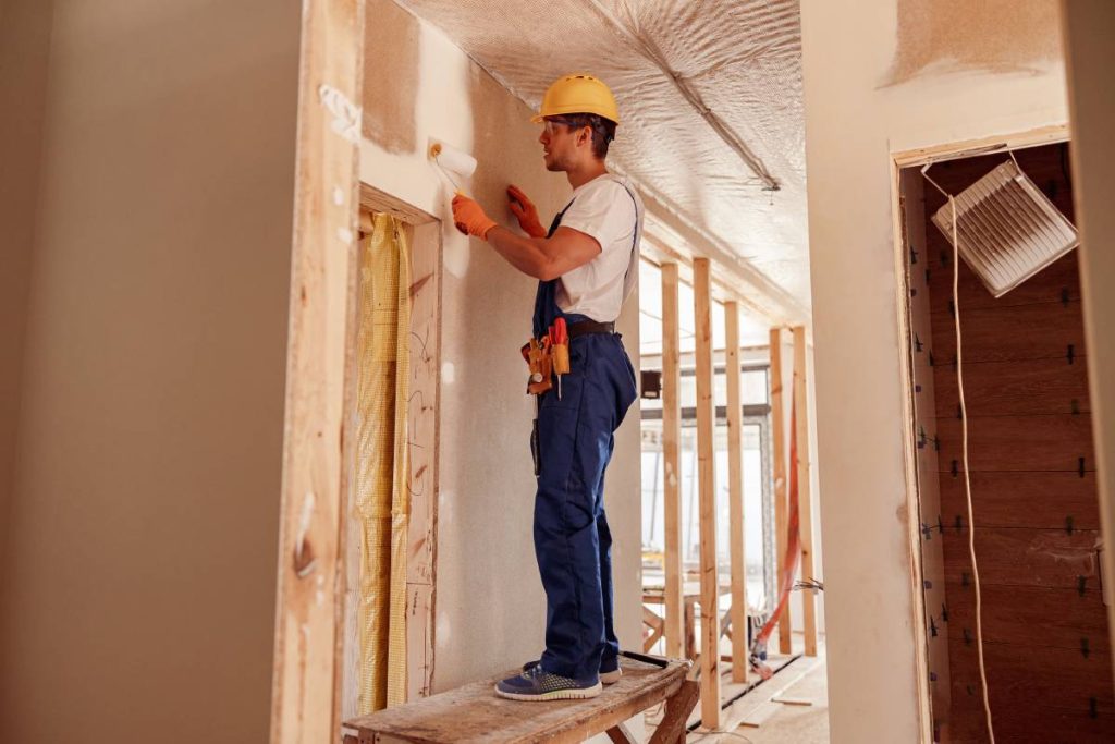 Handsome young man in safety helmet and work overalls painting wall with paint roller